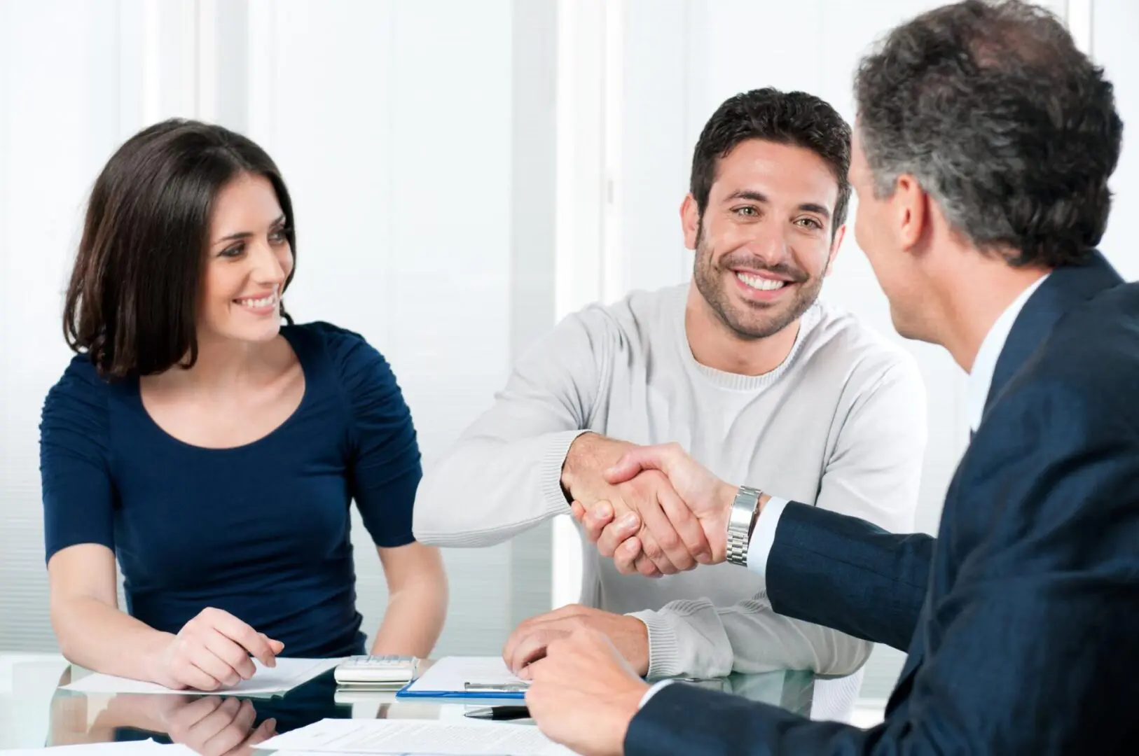 GettyImages-134214747 Three people sitting at a table, with one man shaking hands with another man while a woman watches and smiles. Documents and a calculator are on the table.