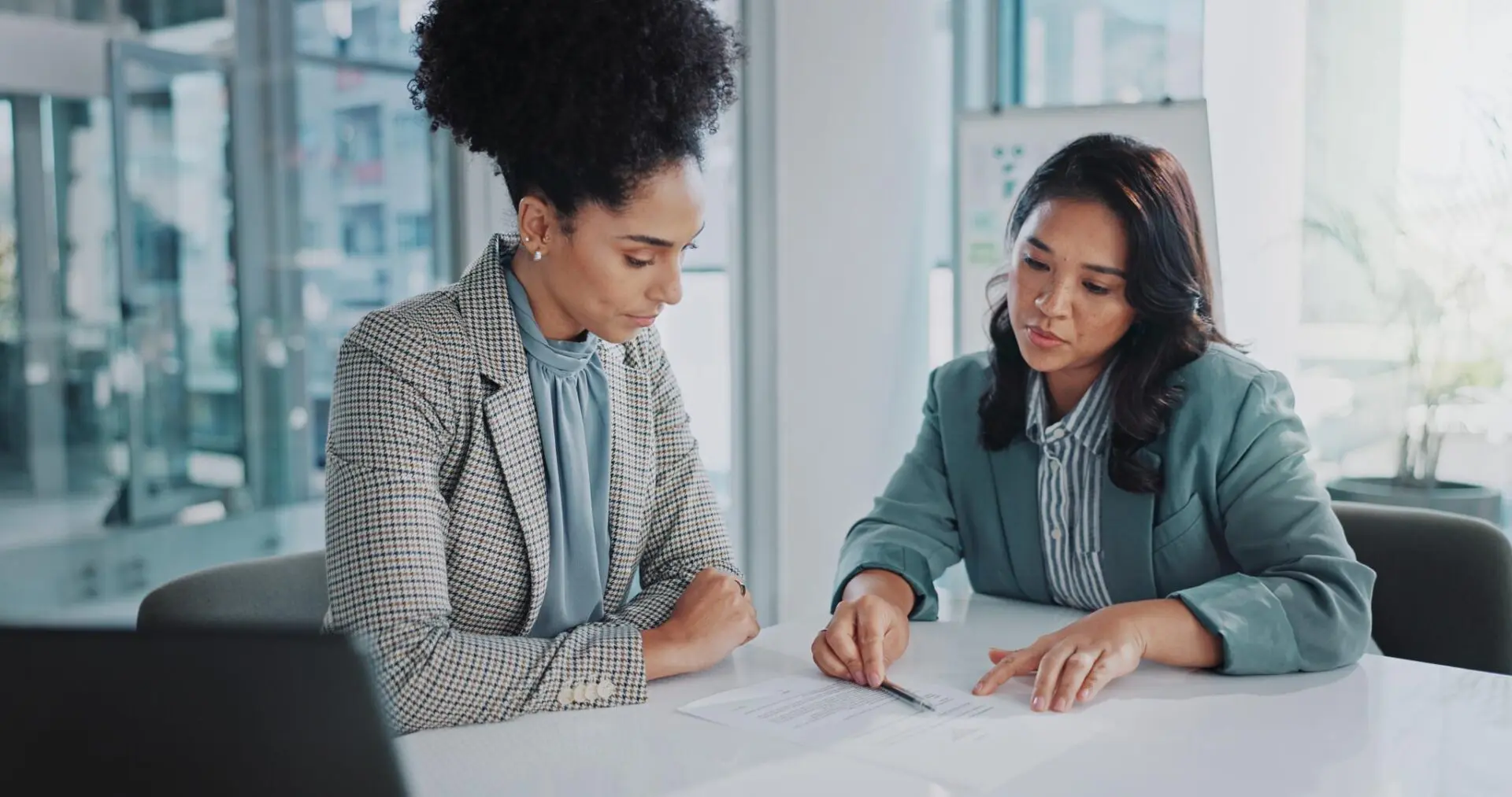 GettyImages-2162643081 Two women in formal attire discussing a document at a table in an office setting.