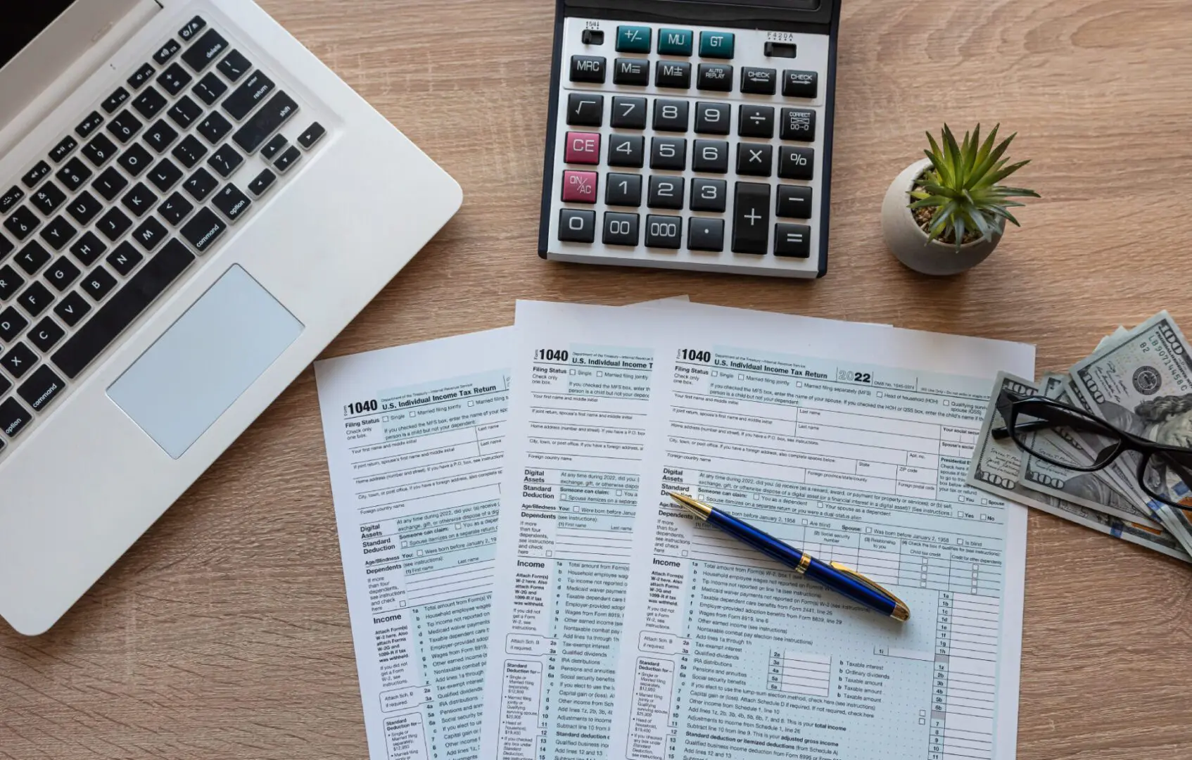GettyImages-546183298 (1) A desk with tax documents, a pen, a calculator, a laptop, a small plant, and stacks of U.S. dollar bills.