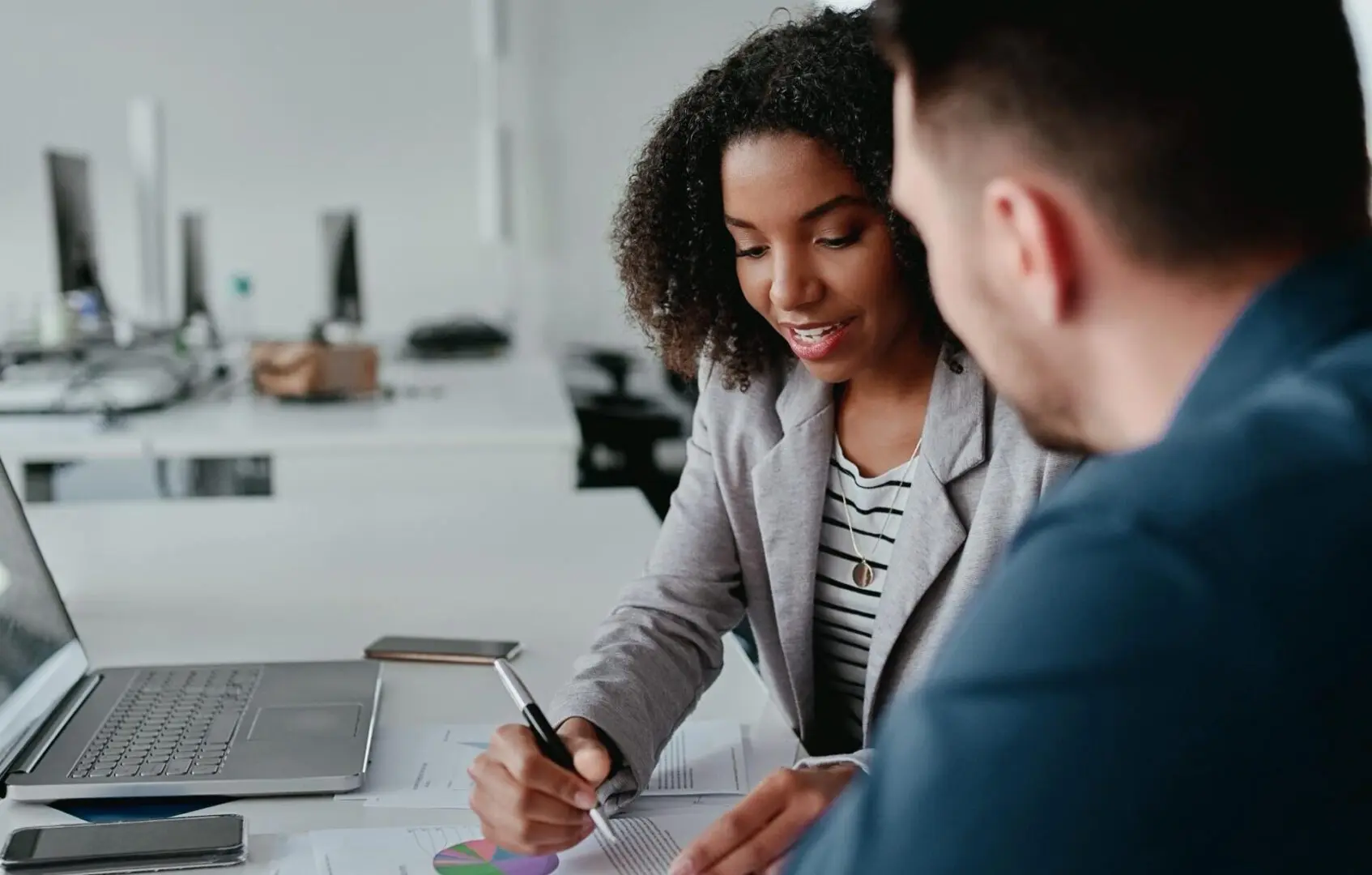 GettyImages-546183298 Two people sit at a desk with a laptop, discussing documents. One person is holding a pen, gesturing towards charts on paper.